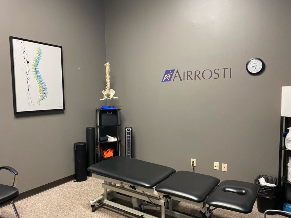 Photo of the treatment room at Airrosti Far Hills. Photo included the Airrosti Logo and the treatment table.
