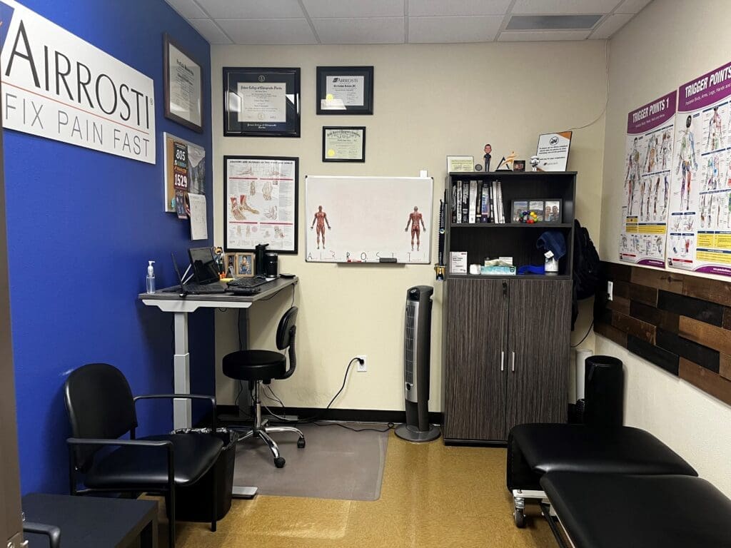 a view of the interior treatment room at Airrosti Forney. The provider's desk and acolades decorate the walls. A treatment table is visible on the right side of the frame.