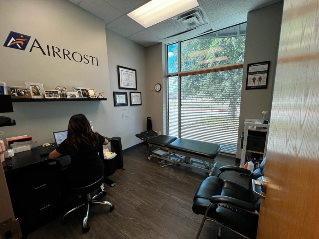 a photo of one of the treatment rooms at Airrosti Rowlett. The Airrosti logo is printed on the wall. A treatment table is visible in the center of the room by the window.