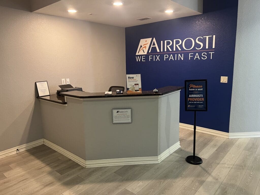 Panned out interior view of the front desk at Airrosti South Fort Worth. Includes the Airrosti logo on blue wall behind the desk
