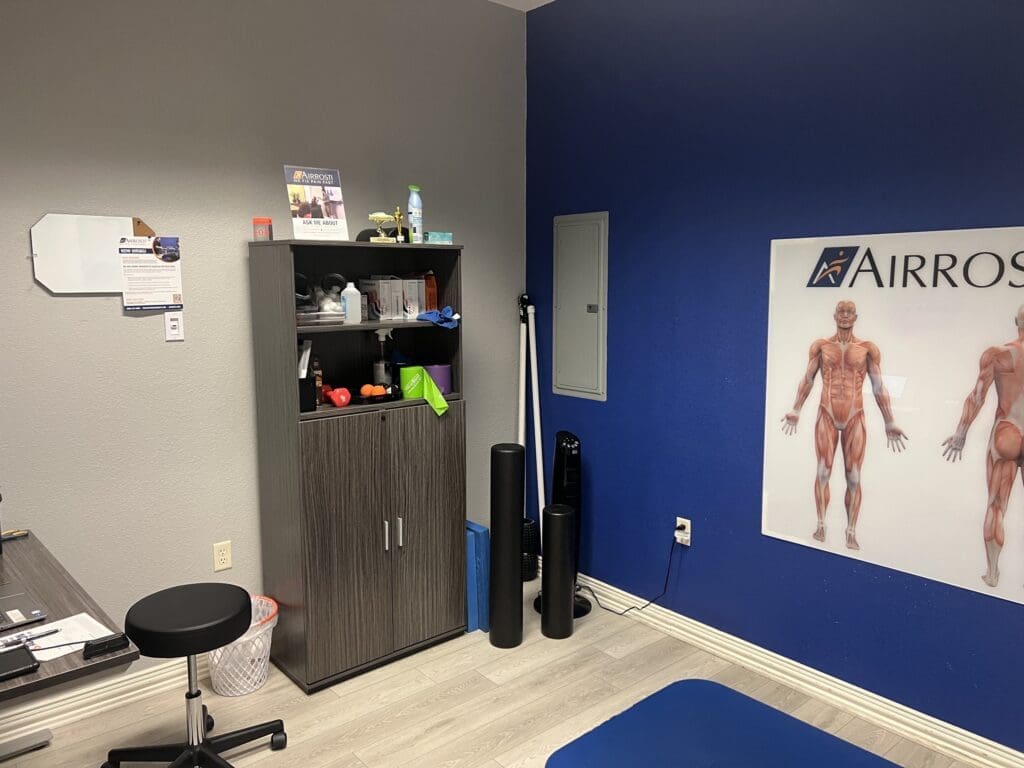 an interior photo of the recovery room at Airrosti South Fort Worth. The exercise mat and musculoskeletal man poster are visible. Includes foam rollers, sticks, and kinesiology tape