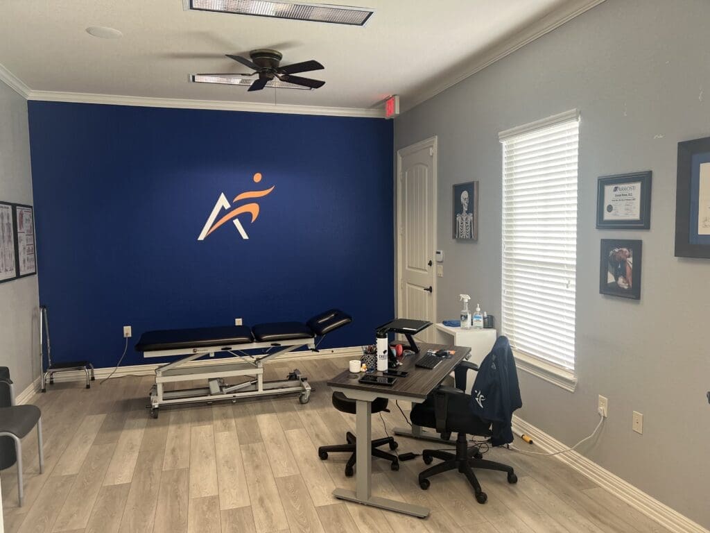 An interior view of the treatment room at Airrosti South Fort Worth. Includes the provider's desk and the treatment table. The Airrosti logo is visible on back wall.